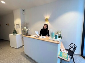 A reception desk with a woman standing behind it, holding a document. There are decorative plants and a small Christmas tree on the counter. - Agora Wellness suites (Tournai)
