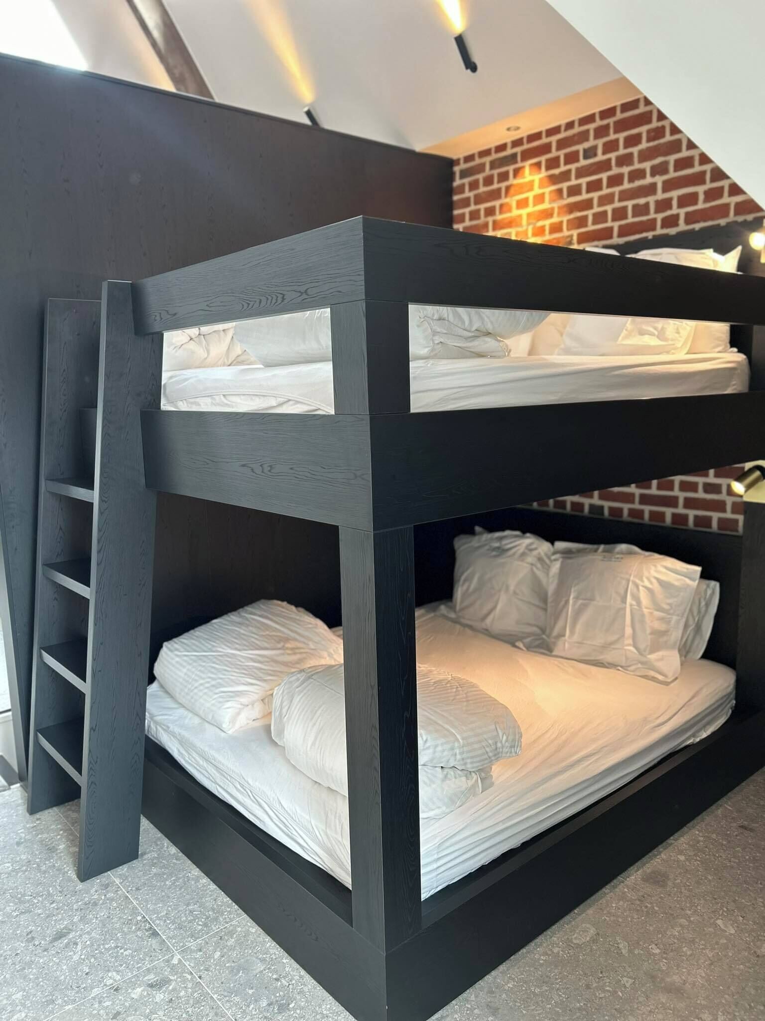 A bunk bed with a ladder, white bedding, and a brick wall in the background.