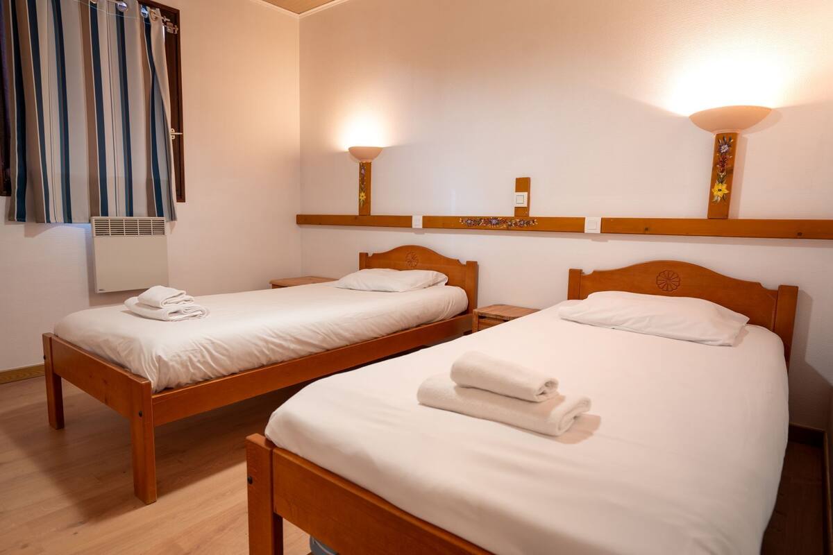 Two single beds with wooden frames and white linens, a radiator, and a wall-mounted shelf with decorative items.