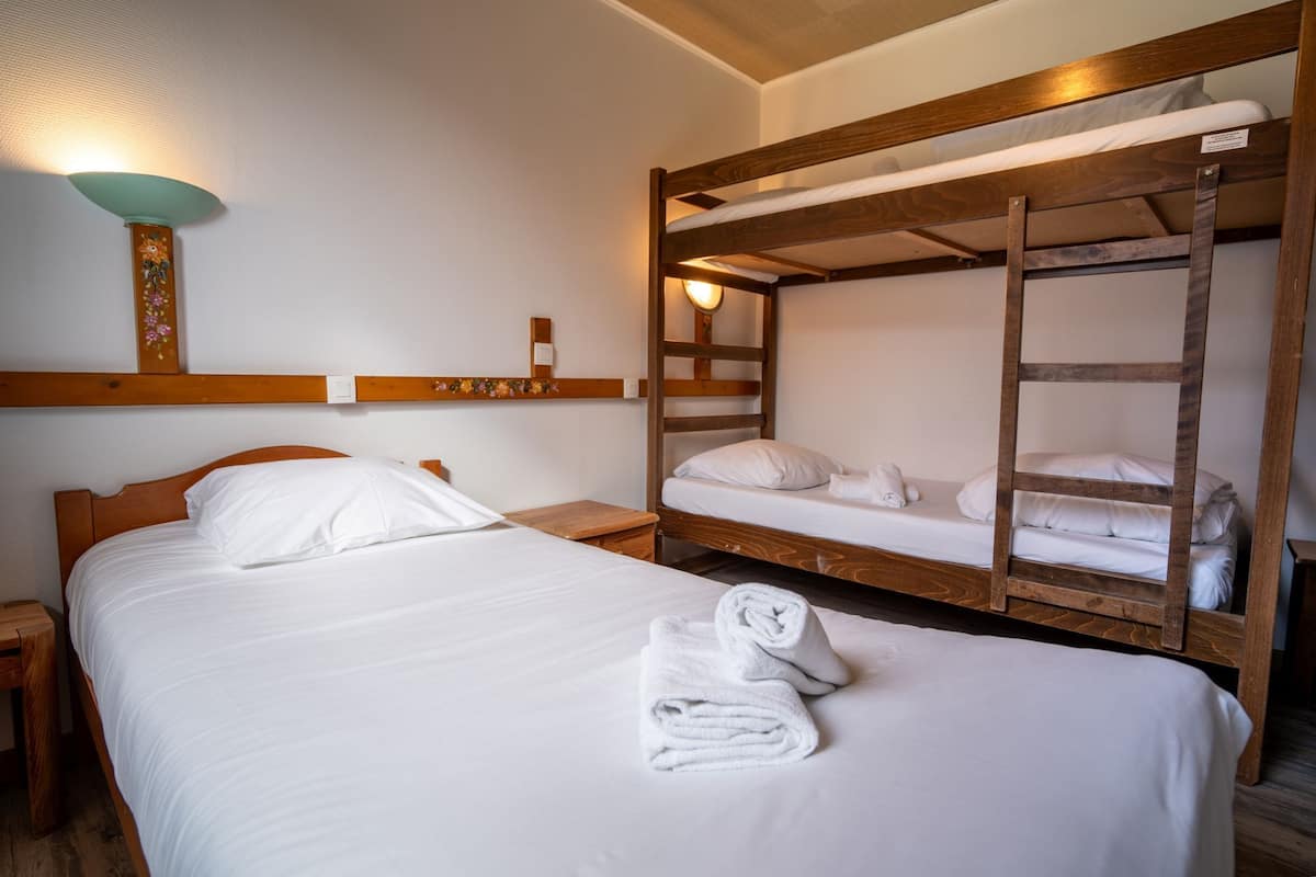 A room with a single bed and two bunk beds, wooden headboards, a wall-mounted light fixture, and a small shelf with a decorative item.