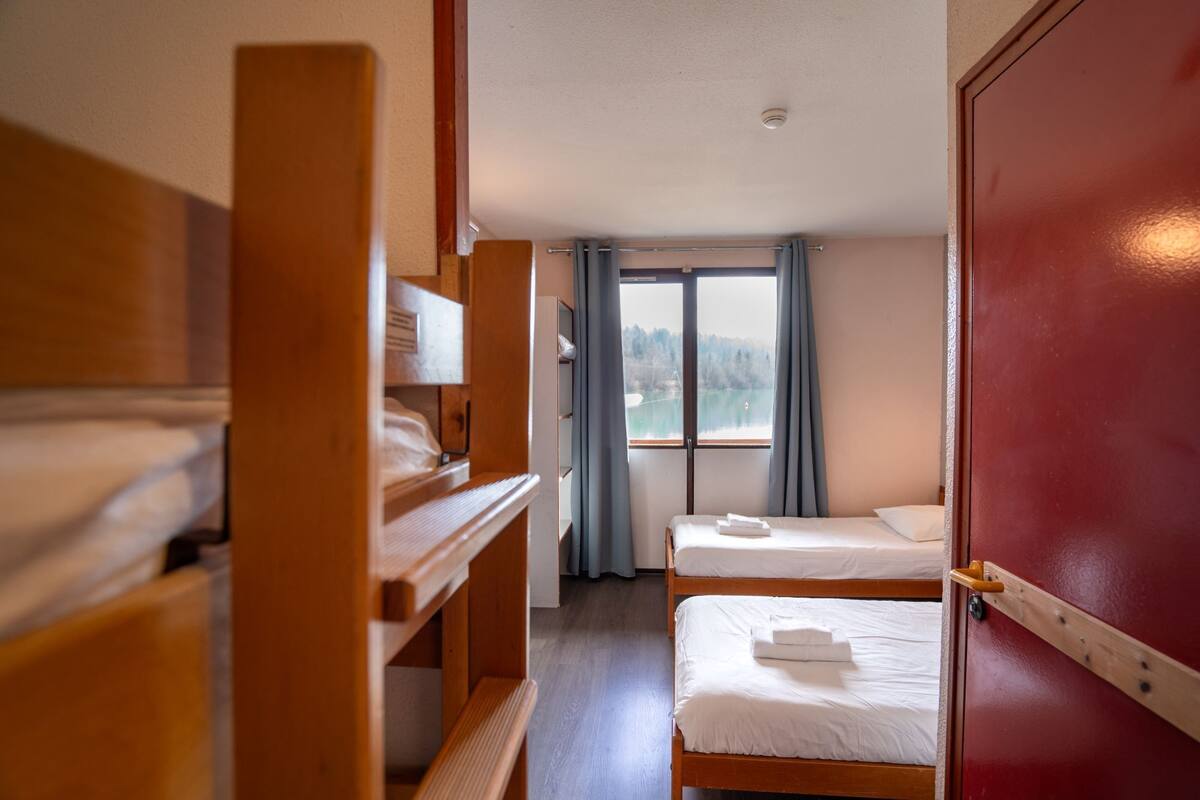 A room with bunk beds, a wooden wardrobe, a window with curtains, and a view of a lake.