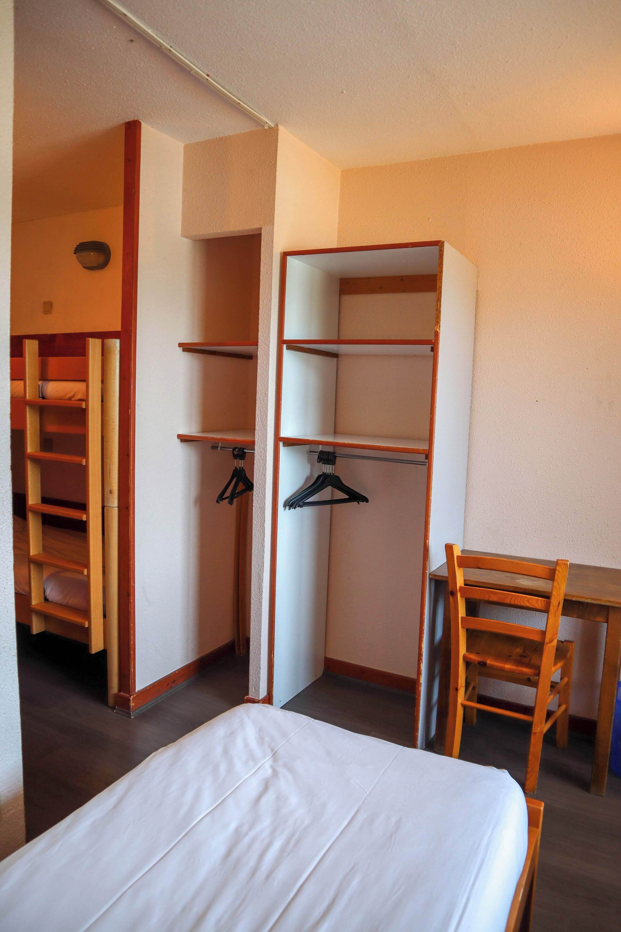Een slaapkamer met een bed, een houten stoel, een bureau en een inbouwkast met planken en hangruimte.