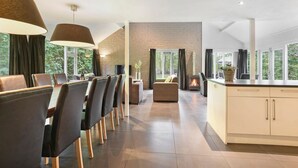 Et moderne spiseområde med et langt bord, stole, en pejs og store vinduer der giver udsigt til grønne omgivelser.
