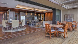 Een ruime binnenruimte met houten vloeren, een zitgedeelte in het midden met ronde tafels en stoelen, en een keuken die op de achtergrond zichtbaar is.
