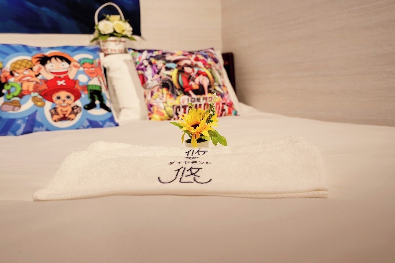Ein ordentlich bezogenes Bett mit einem weißen Handtuch und japanischen Schriftzeichen, eine kleine Vase mit einer gelben Blume und farbenfrohe Kissen mit Zeichentrickfiguren.