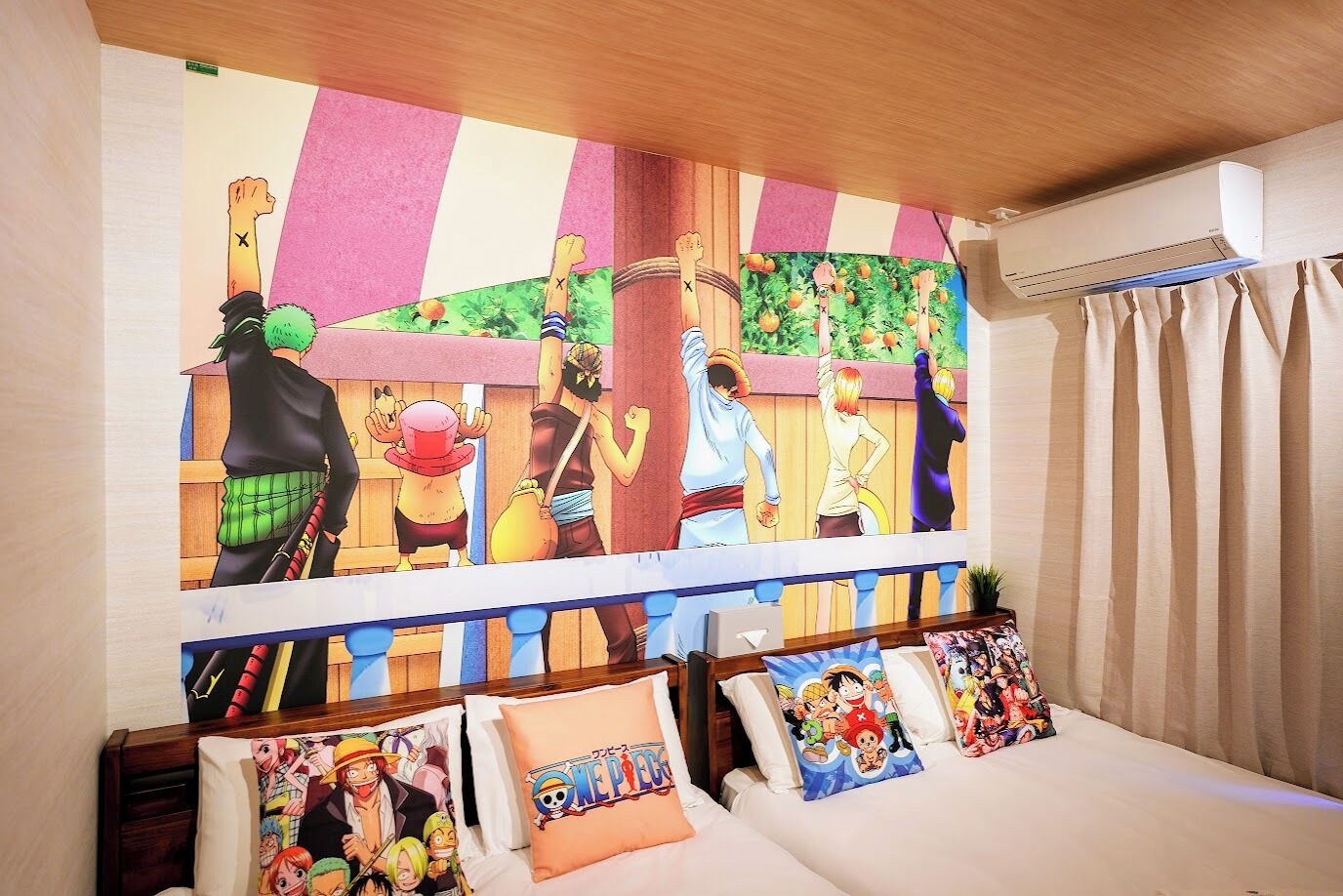 A hotel room with a large anime-themed mural on the wall, a bed with multiple pillows featuring anime characters, and a window with curtains.
