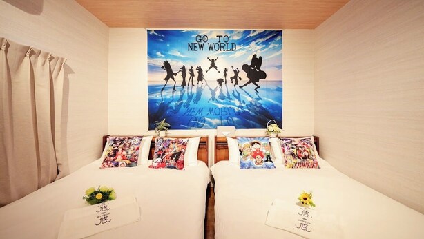 Habitación de hotel con dos camas, cada una con un cojín que presenta personajes de anime. Un cartel grande en la pared dice "GO TO NEW WORLD".