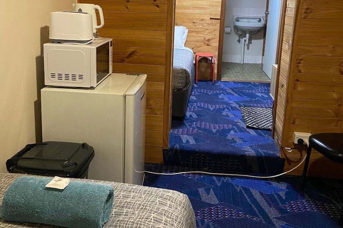 Quarto compacto com cama, micro-ondas, uma pequena mesa e uma cadeira.
