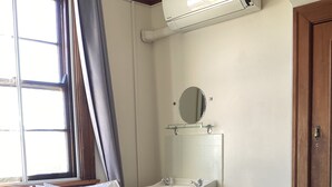 Un dormitorio con cama, ventana con cortinas, aire acondicionado de pared, lavabo y espejo.