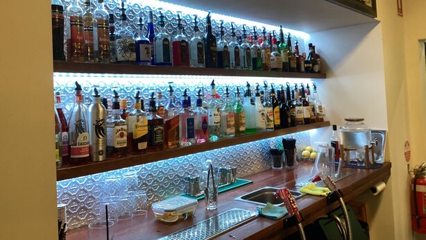 Zona de bar con estantes llenos de botellas de licores, un lavamanos y un extintor.