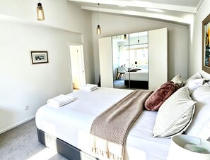 A modern bedroom with a large bed, a mirror, and a view into an adjacent room.