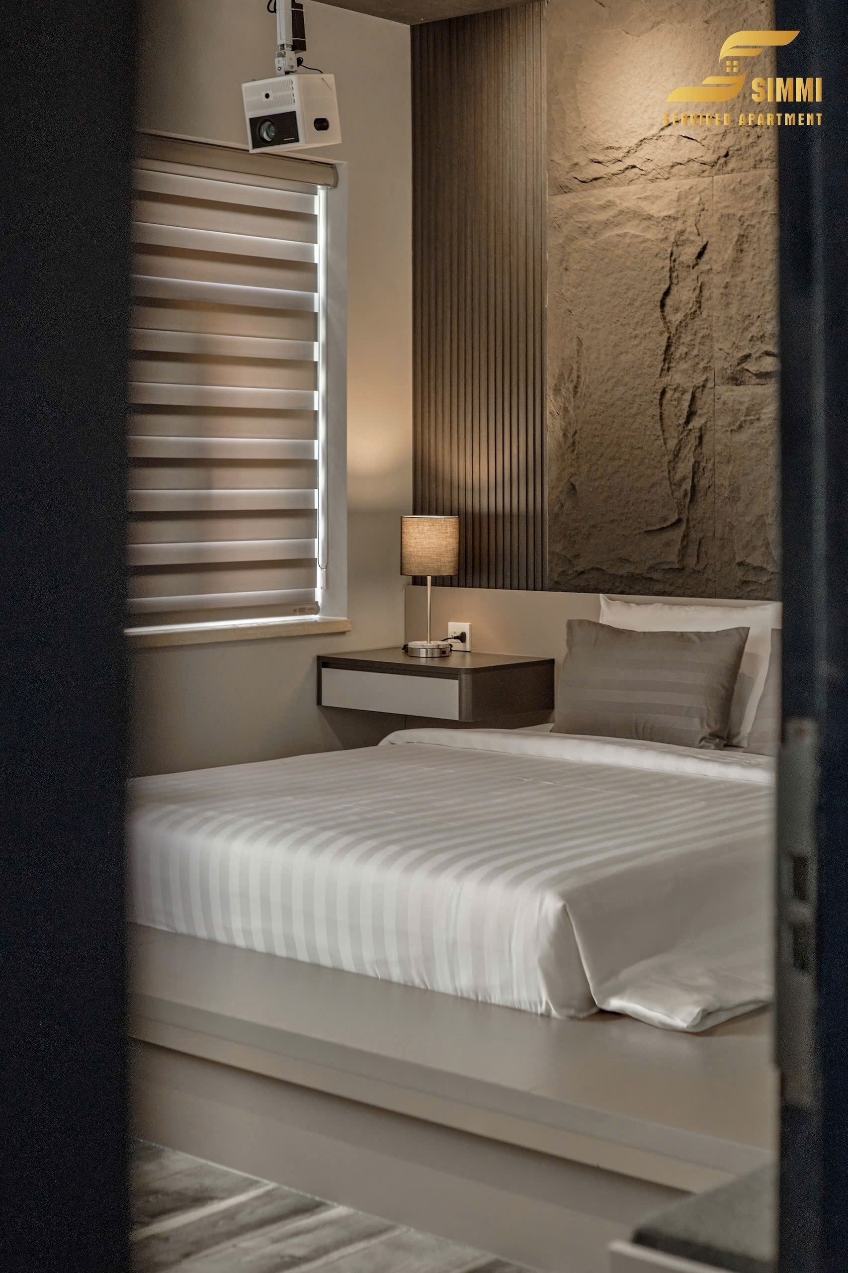 A modern bedroom with a bed, bedside table, lamp, and a wall-mounted projector.