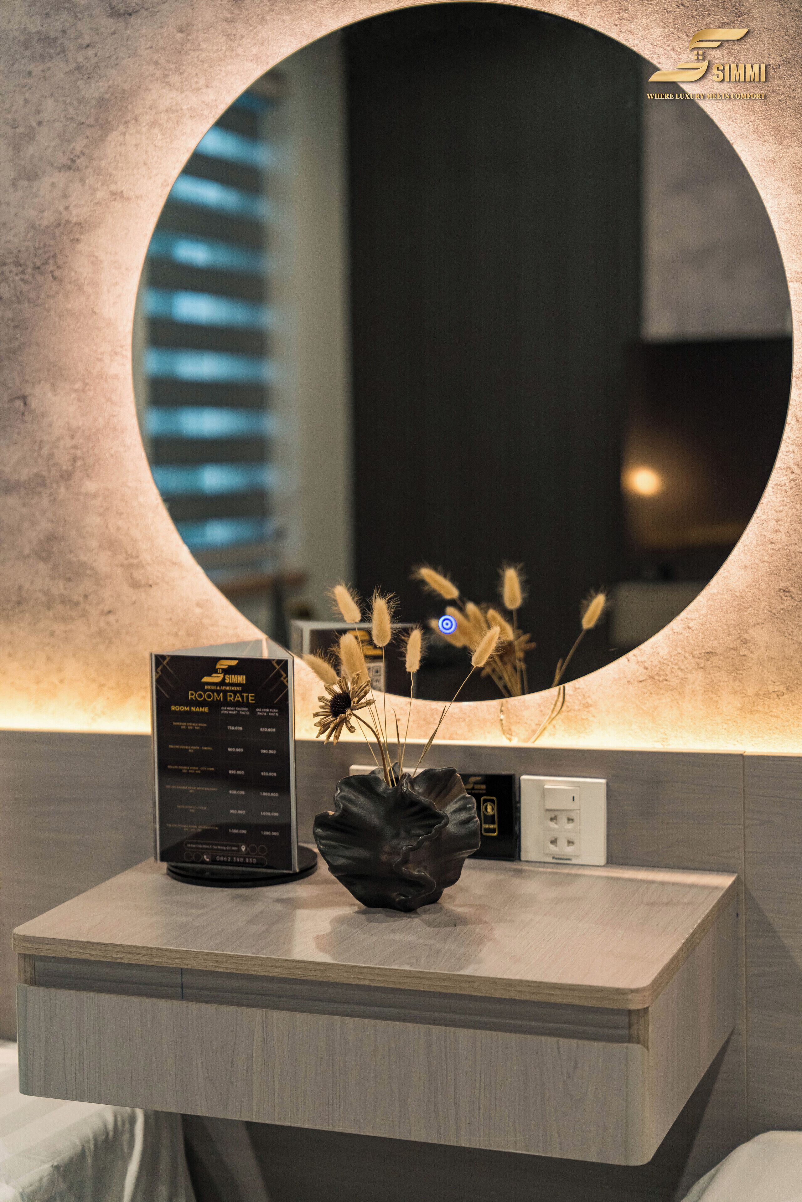 A modern hotel room with a round mirror, a black digital display for room rates, a vase with dried flowers, and a power outlet on the wall.