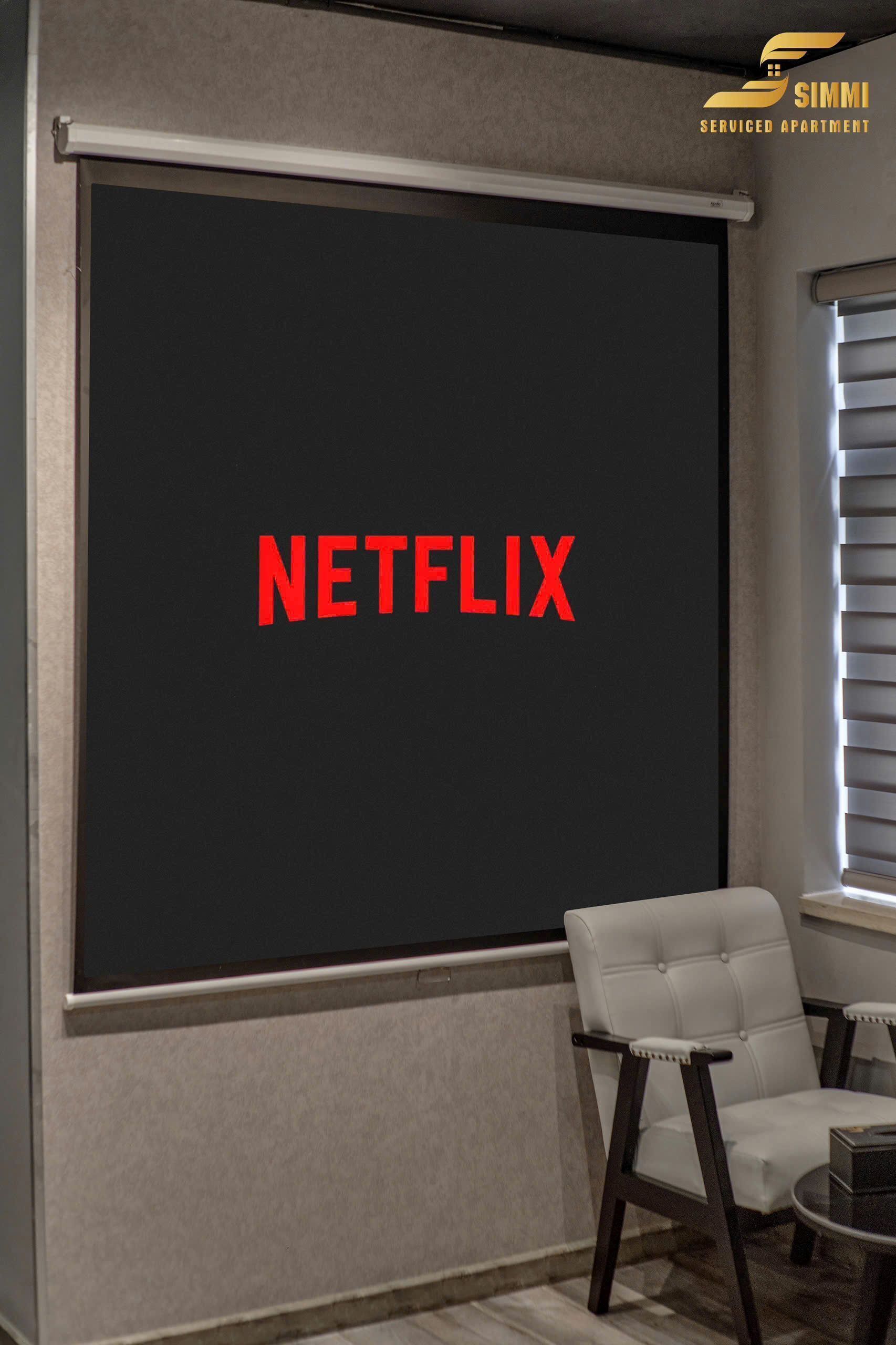 A room with a large black screen displaying "NETFLIX" in red letters, a white chair, and a small table.