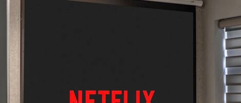 A room with a large black screen displaying "NETFLIX" in red letters, a white chair, and a small table.