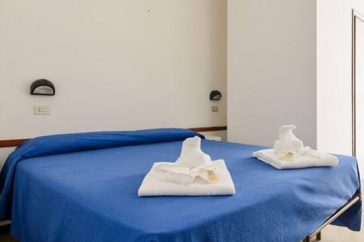 A hotel room with a single bed, a blue bedspread, and two white towels folded on the bed.
