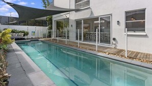 A modern outdoor swimming pool with a wooden deck, glass doors, and a shaded area. - The Seabreeze Sanctuary (WOORIM)