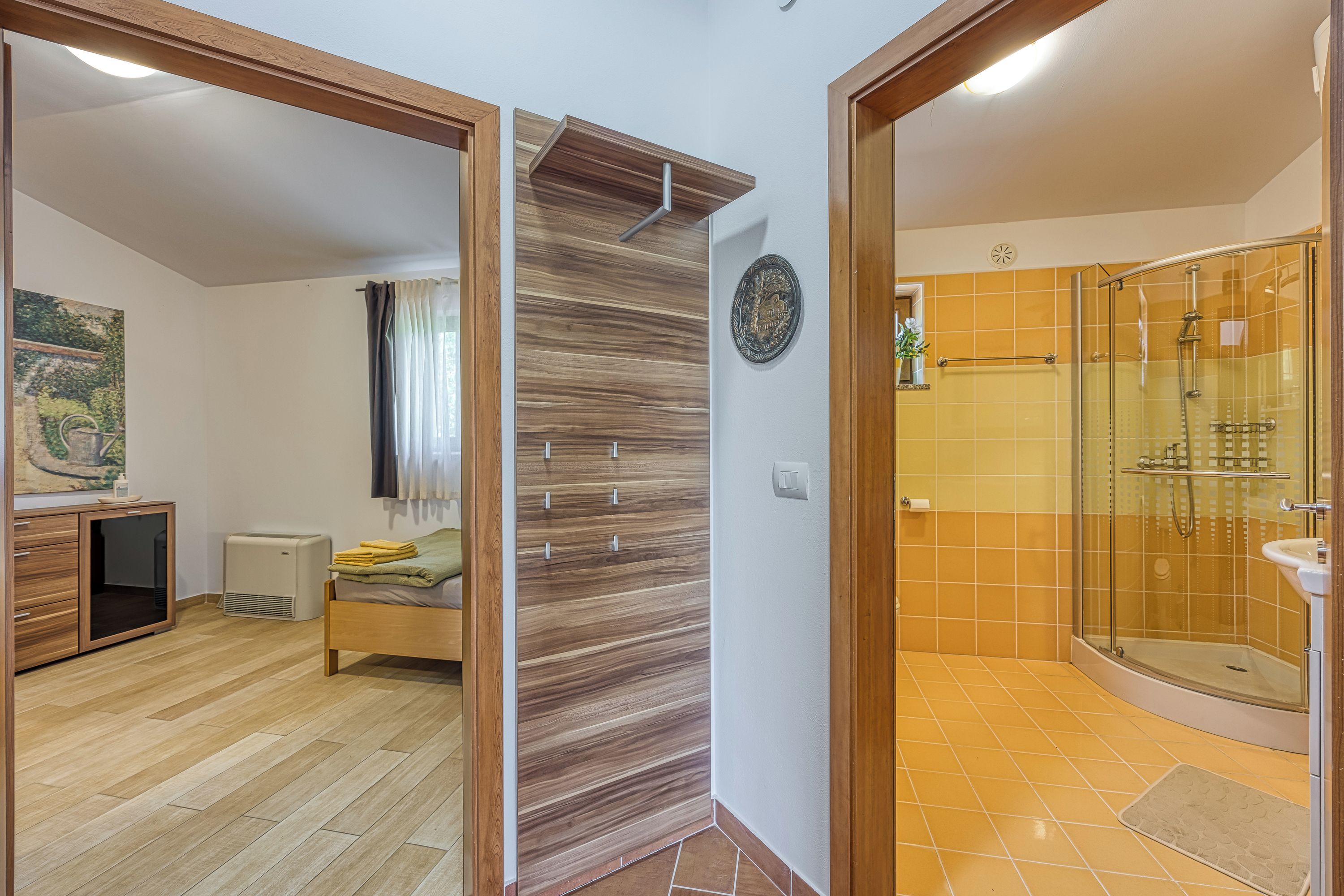 A modern bathroom with a glass shower enclosure, a wooden floor, and a bed with a green blanket.
