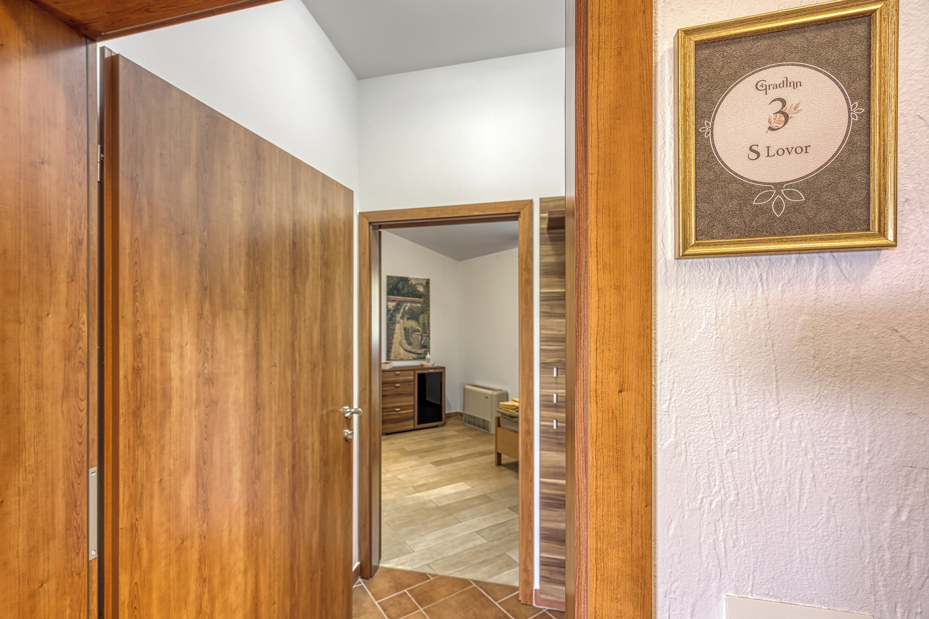 A wooden door leading to a room with a tiled floor, a framed certificate on the wall, and a small desk with a chair.