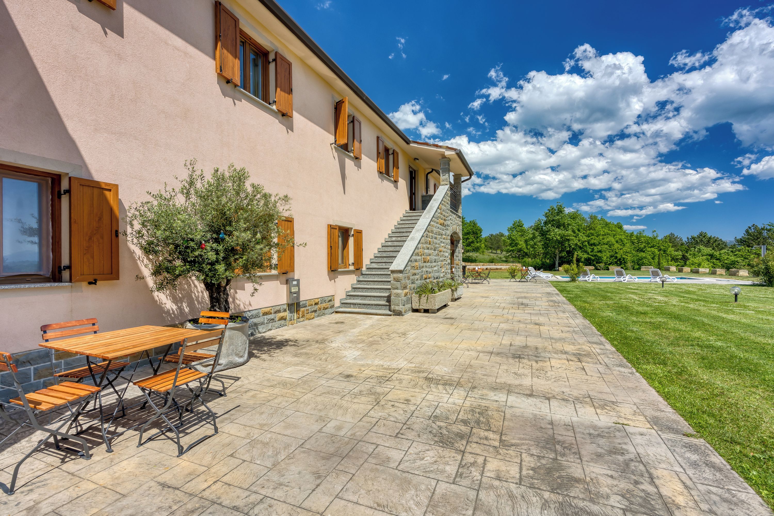 A building with a stone staircase leading to a second floor, outdoor seating with tables and chairs, and a well-maintained lawn.
