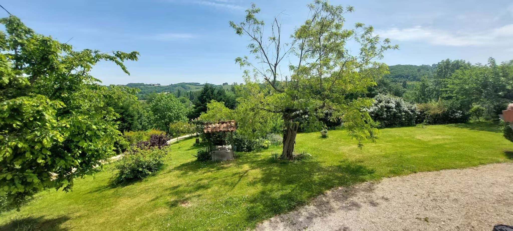 Een verzorgde tuin met diverse bomen en struiken, een grindpad en een helder uitzicht over het omliggende landschap.