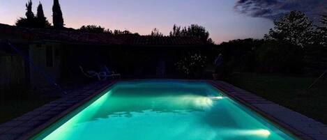 Une piscine rectangulaire dont le bord est recouvert de carreaux, entourée d’un petit muret en briques. Des chaises longues sont disposées le long du muret, et la piscine est éclairée par une lumière verte.