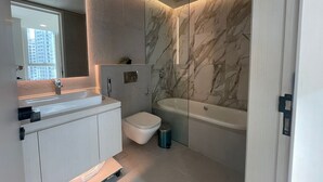 A modern bathroom with a marble wall, a bathtub, a toilet, and a vanity with a mirror.