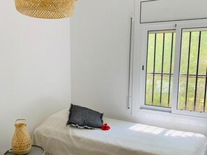 Una habitación con cama, ventana con rejas, lámpara colgante y una pequeña mesa con un objeto decorativo.