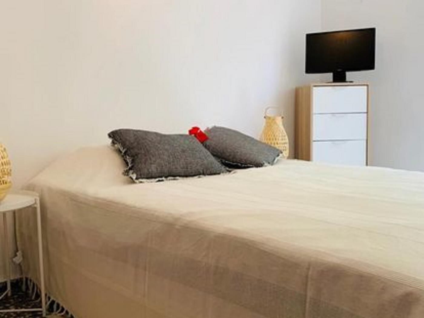 A neatly made bed with a white bedspread and two gray pillows, a small white table with a lamp, and a TV on a stand against the wall.