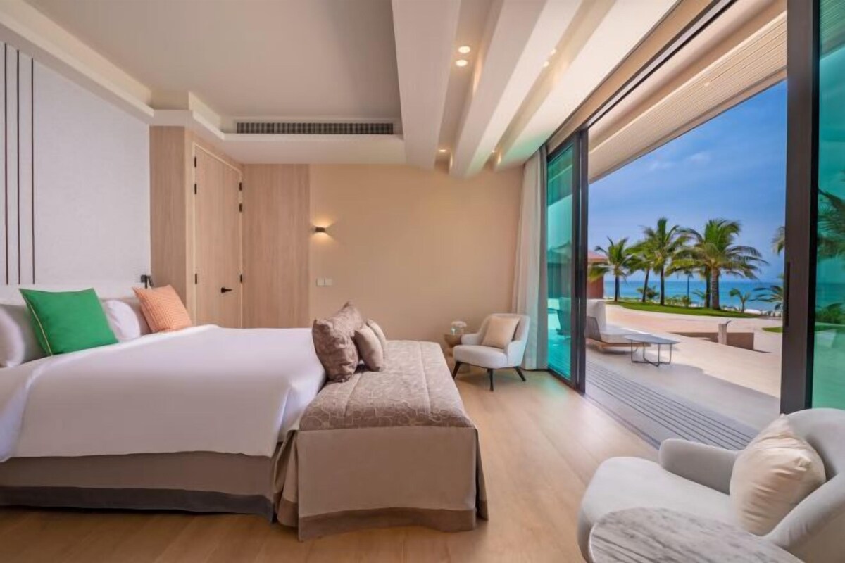 A modern hotel room with a large bed, a seating area, and a balcony with a view of palm trees and the ocean.