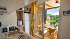 A compact kitchen with a gas stove, sink, and a refrigerator. It features a sliding glass door leading to an outdoor wooden deck with a table and chairs. - Lastura Mobile Homes (Novalja)