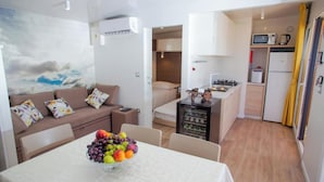 A compact living space with a kitchenette, a dining area with a fruit bowl, a sofa with floral cushions, and a painting of clouds on the wall. - Lastura Mobile Homes (Novalja)