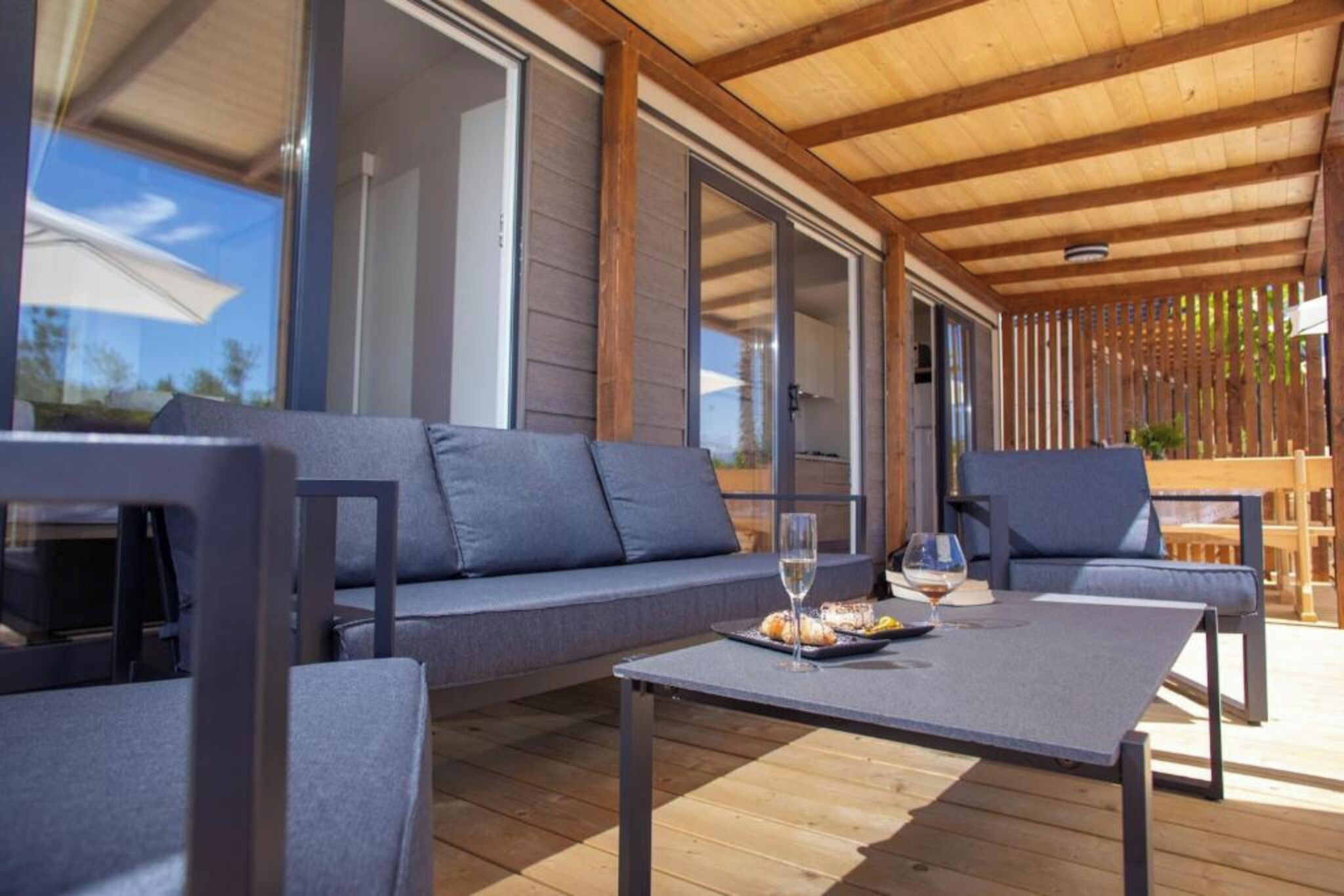A covered outdoor seating area with a wooden ceiling, grey sofas, and a table with a glass of wine.