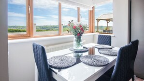 Une table à manger avec quatre chaises, un vase rempli de fleurs et une vue sur la campagne, visible à travers de grandes fenêtres.