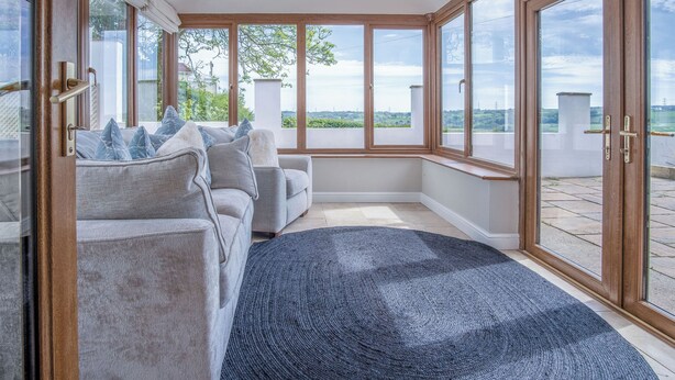 Un salon moderne avec un canapé gris, des coussins bleus et une grande fenêtre donnant sur un paysage verdoyant et des bâtiments.