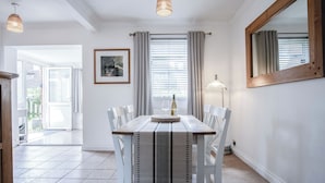 A dining area with a table set for two, white chairs, a woven table runner, a framed picture on the wall, and a window with curtains. - Sunderland Avenue - 2 Bedroom - Pembroke Dock (Pembroke Dock)