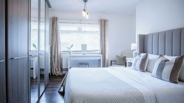 A modern hotel room with a large bed, a desk, and a view through a window with curtains. - Sunderland Avenue - 2 Bedroom - Pembroke Dock (Pembroke Dock)