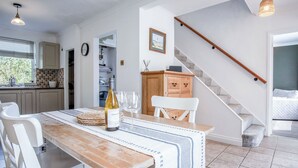 A dining area with a wooden table, white chairs, and a bottle of wine. A staircase is visible in the background. - Sunderland Avenue - 2 Bedroom - Pembroke Dock (Pembroke Dock)