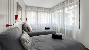 Une chambre d’hôtel avec deux lits, une lampe murale rouge, des rideaux légers et une vue sur la ville.