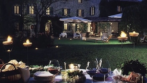 An outdoor dining setup with tables, chairs, and candles, set against a stone building with a garden. - La Magioca (Negrar)