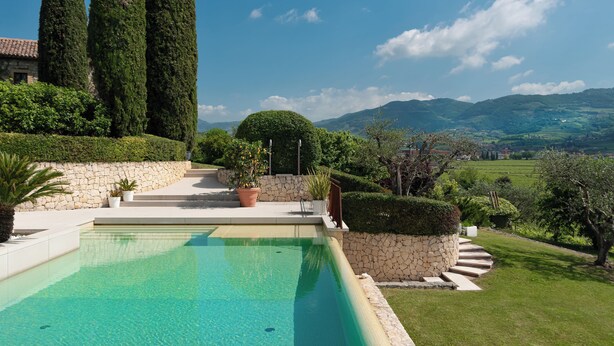A swimming pool with a stone wall and steps leading to a garden with a view of distant hills. - La Magioca (Negrar)