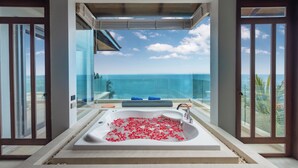 A modern bathroom with a large bathtub filled with rose petals, a glass-enclosed shower, and a view of the ocean. - Villa La Thalé (Kamala)