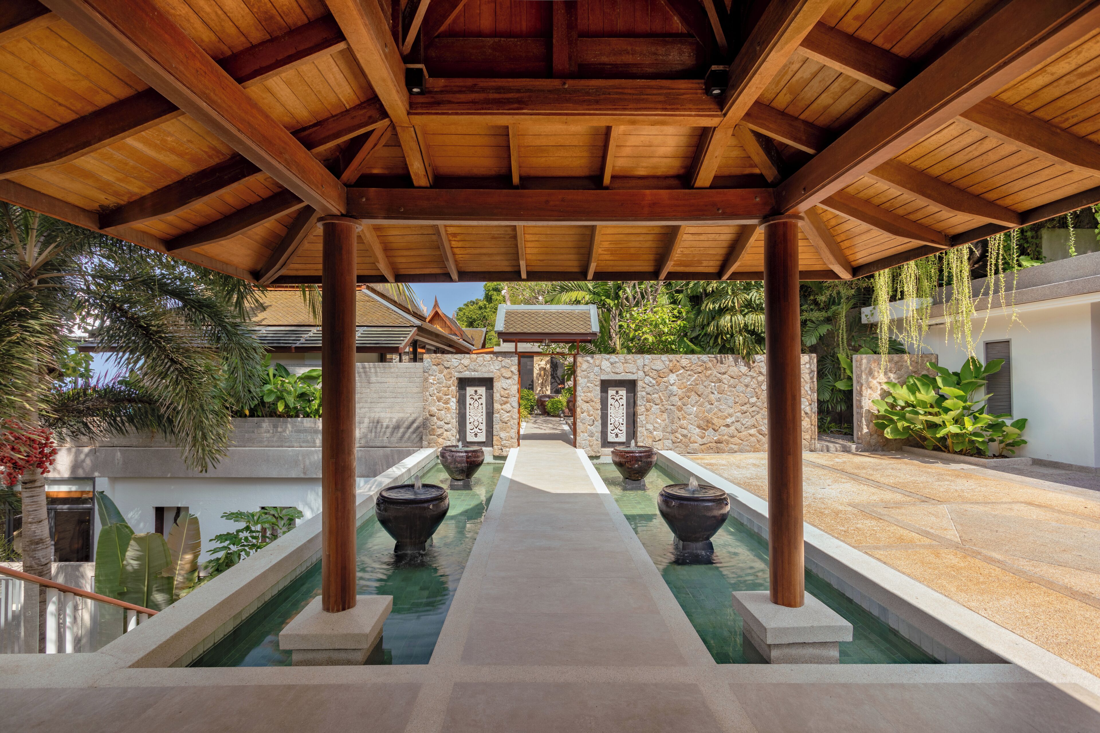 A covered pool area with a wooden ceiling, stone walls, and a pathway leading to a building entrance.