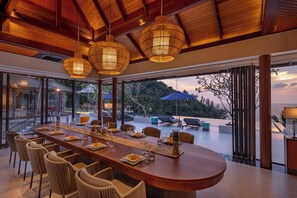A spacious dining area with a long wooden table set for a meal, surrounded by comfortable chairs, and large windows offering a view of the outdoors. - Villa La Thalé (Kamala)
