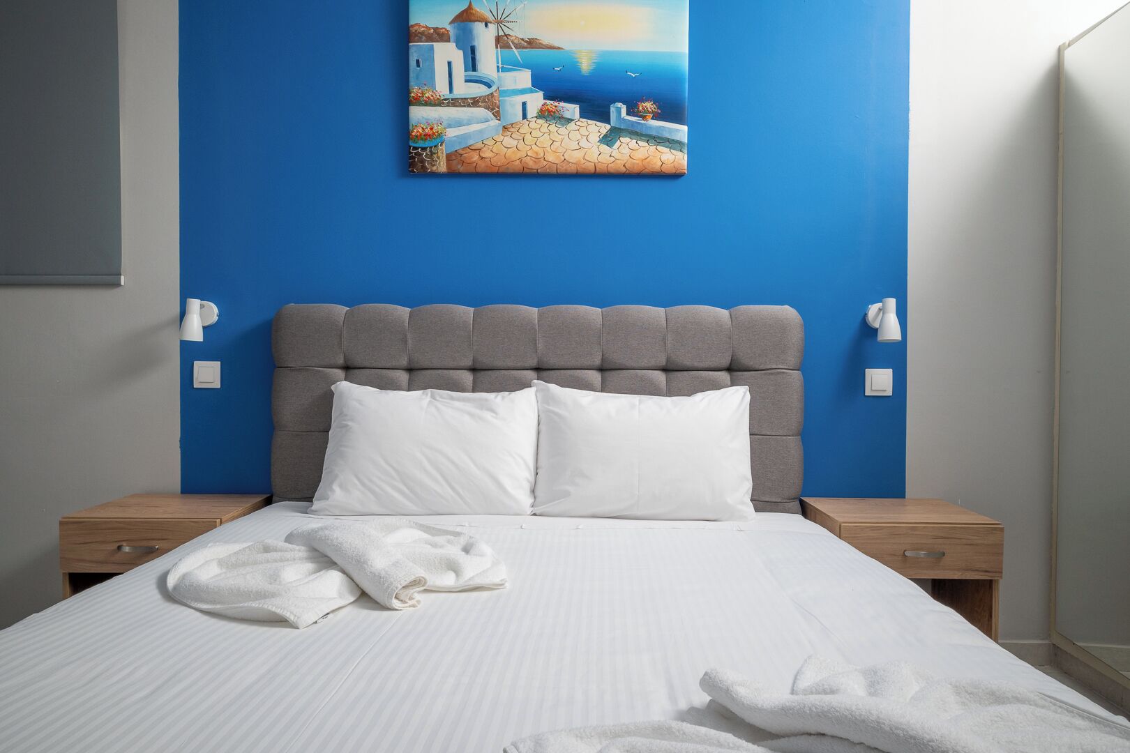 A neatly made bed with white linens and two pillows, a grey upholstered headboard, and a painting of a coastal scene above the bed.