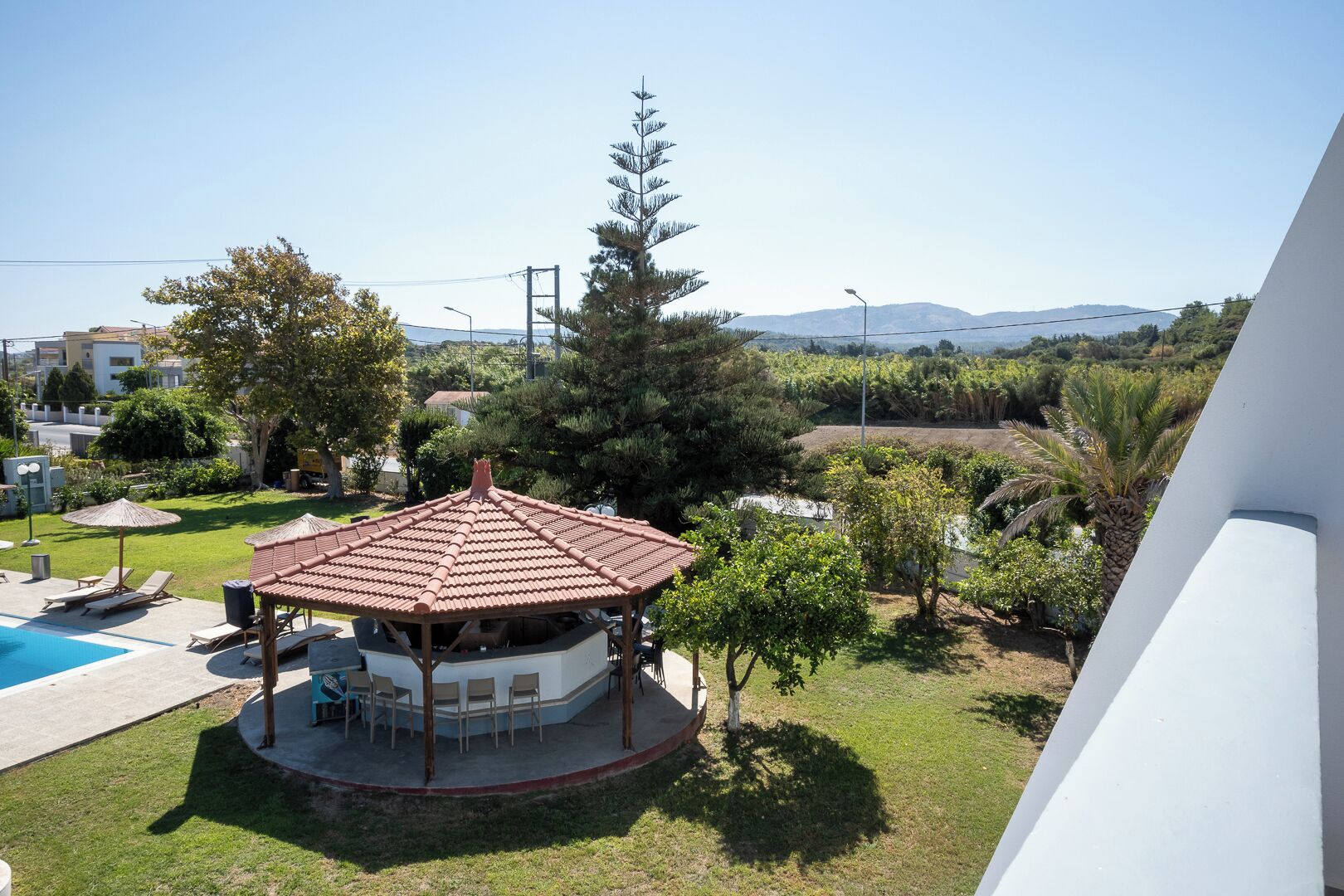 A gazebo with a tiled roof overlooking a swimming pool, surrounded by greenery and a mountainous backdrop.