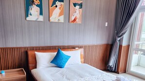 Een net opgemaakt bed met een witte quilt en blauwe accenten, een houten nachtkastje met een lamp, en drie kunstwerken aan de muur erboven.