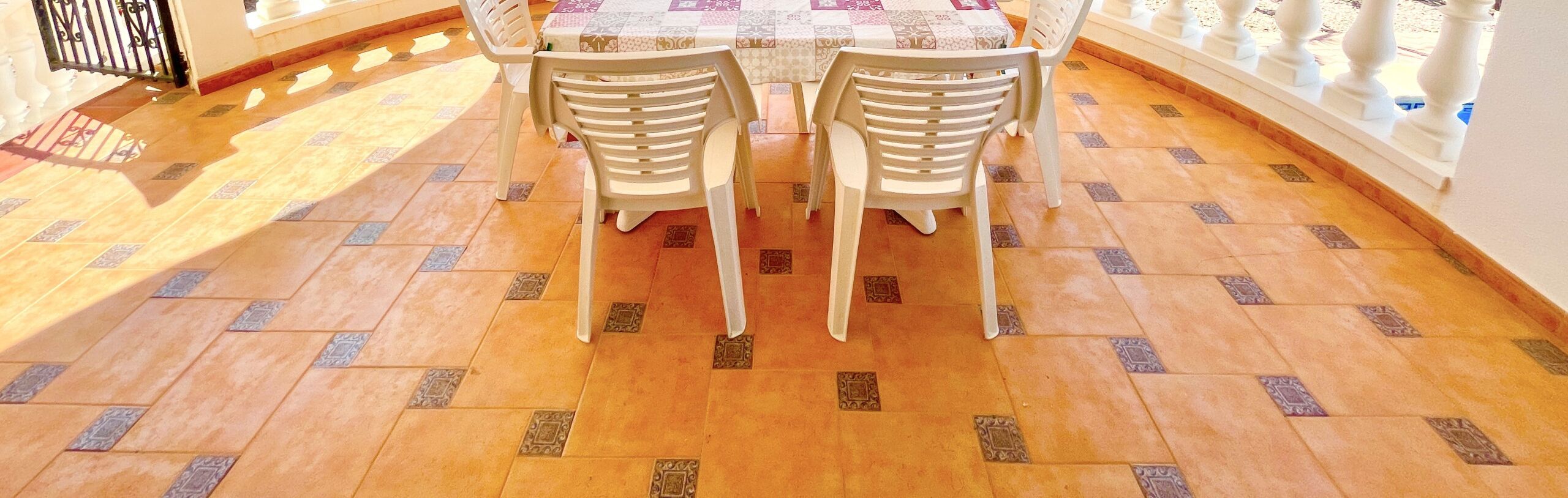 Un comedor con una mesa cubierta por un mantel estampado, dos sillas de plástico blanco y un piso de baldosas con un diseño geométrico.
