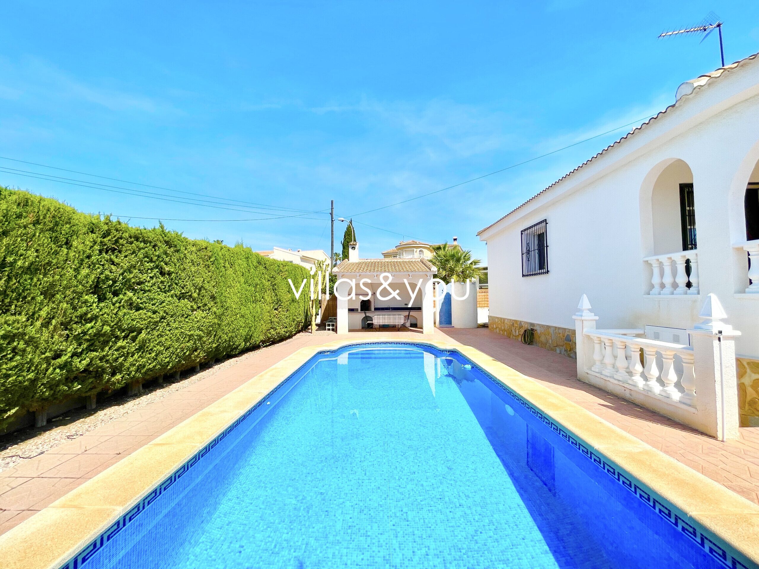 Piscina com borda azulejada, cercada por um edifício branco com arcos e um muro branco.