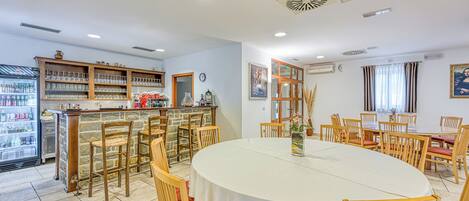 A restaurant with a round table, wooden chairs, a bar area, and a refrigerator stocked with drinks.